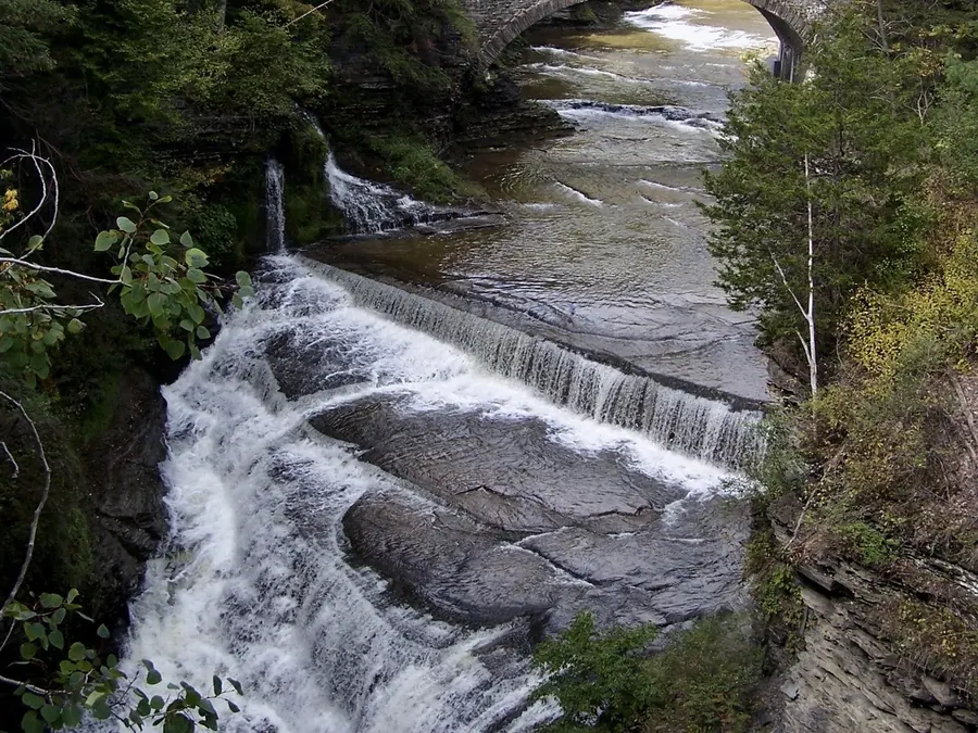 A waterfall pouring over rock ledges beneath a stone arch bridge in a wooded gorge.